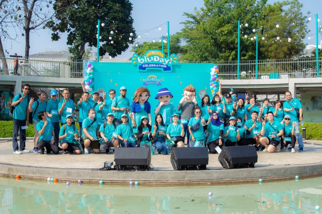 Foto grup besar dengan maskot dan backdrop bluDay Celebration di siang hari